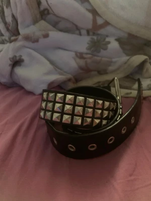 Svart nitar bälte med metallspänne - Säljer ett svart bälte med stora silverfärgade nitar och metallspänne. Bältet har flera hål med metallringar och en bred design som ger en edgy look. Perfekt accessoar för att lyfta din outfit.