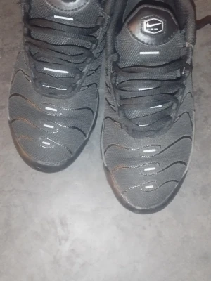 Nike Air Max Plus svarta sneakers - Säljer ett par svarta Nike Air Max Plus sneakers med klassiska vågiga detaljer och mesh-överdel. Skorna har svarta skosnören, diskret Swoosh på sidan och en ikonisk Air-logga på plösen. Perfekta för dig som gillar streetwear och sportig stil. Vet ej om de är äkta 