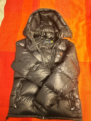 Glossy Svart pufferjacka från Polo Ralph Lauren - Säljer en svart dunjacka från Polo Ralph Lauren med huva och dragkedjor på fickorna. Jackan har en klassisk puffer-modell och den ikoniska röda loggan broderad på bröstet. Perfekt för kalla vinterdagar och riktigt skön att bära. Handledsremmen är svag, men den kan lätt fixas.