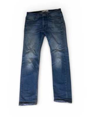 Blå raka jeans med slitningar - Säljer ett par blå jeans med raka ben och snygga slitningar vid knäna. Jeansen har klassisk femficksdesign och en tvättad look som ger en avslappnad vibe. Perfekta för dig som gillar en casual stil. Materialet är bomull.