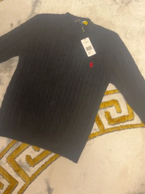 Svart kabelstickad tröja Polo Ralph Lauren - Kabelstickad svart tröja från Polo Ralph Lauren med rund halsringning och den klassiska röda logotypen broderad på bröstet. Tröjan har långa ärmar och ribbade muddar vid ärmslut och nederkant. Perfekt för dig som gillar stilrena och tidlösa plagg.