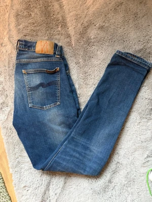 Blå Nudie jeans w30 l32 - Säljer ett par klassiska blå jeans från Nudie Jeans med snygga slitningar. Storlek W30 L32. Jag är 180 cm lång och de sitter fint. Modellen är tight terry. Hör av er vid minsta fundering!