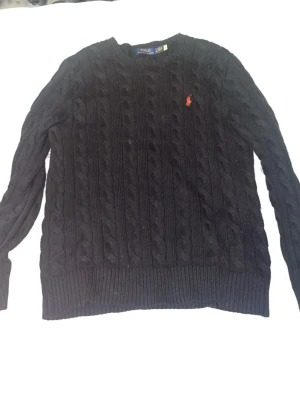 Svart kabelstickad tröja Polo Ralph Lauren - Svart kabelstickad tröja från Polo Ralph Lauren med rund halsringning och röd broderad logga på bröstet. Tröjan har ribbstickade muddar vid ärmslut och nederkant. Perfekt för dig som gillar klassisk och stilren design.