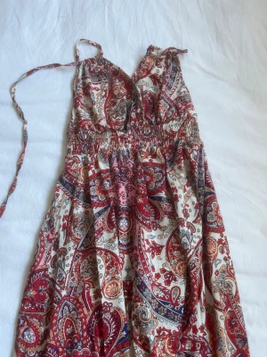 Mönstrad omlottklänning med knyt - Säljer en bohemisk omlottklänning med knytband i nacken och v-ringning. Klänningen har ett färgstarkt paisleymönster i rött, beige och blått samt smock i midjan för en bekväm passform. Perfekt maxiklänning för varma dagar och sommarens alla äventyr.