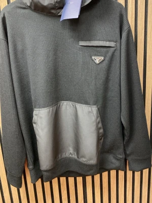 Svart hoodie  med fickor - Snygg svart hoodie  med ribbad struktur, huva och stora fickor framtill i ett glansigt material och en dekorativ ficka. Perfekt för en trendig och avslappnad stil.