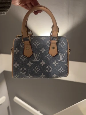 Louis Vuitton blå monogram handväska - Säljer en liten blå handväska från Louis Vuitton i denim-look med klassiskt monogrammönster. Väskan har ljusbruna läderhandtag och guldfärgade metalldetaljer. Perfekt storlek för det viktigaste och en ikonisk accessoar för dig som gillar designer. OBS priset är av att Axelräms bandet är av men går att gå med 