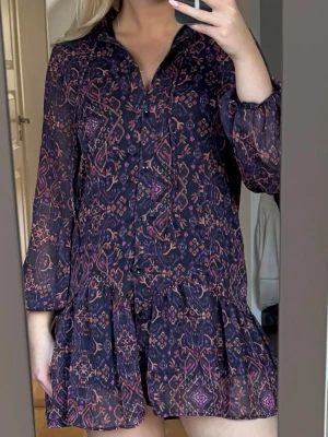 Mörkblå kortklänning med mönster - Kortklänning i mörkblått med lila och röda inslag, har ett bohemiskt mönster och knappar framtill. Klänningen har volang nertill, v-ringning och långa ärmar med lätt puff. Tillverkad i ett tunt, lätt material som ger en luftig känsla🫶🏼