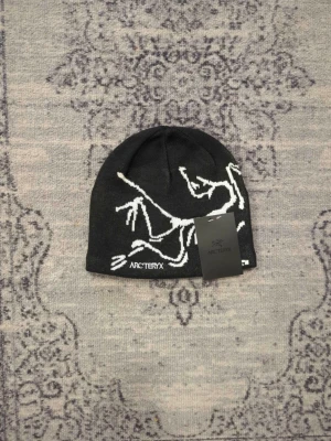 Arc'teryx svart mössa i ull - Snygg svart mössa från Arc'teryx med vit logga och grafiskt skelettmönster. Tillverkad i mjukt, stretchigt material som sitter skönt på huvudet. Perfekt för dig som vill ha en stilren och sportig look.