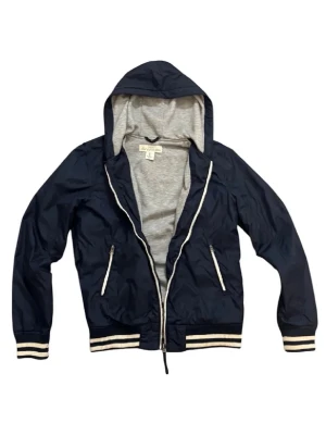 Mörkblå vindjacka med huva - Snygg mörkblå vindjacka med grått foder och huva. Jackan har dragkedja och ribbade muddar i vitt och blått. Perfekt för blåsiga dagar. Har ett litet hål men de syns inte så mycket den är perfekt till våren eller kalla sommar dagar 