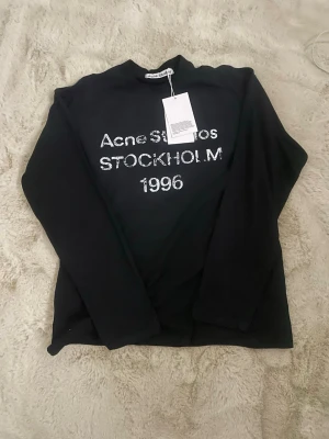 Svart tröja från Acne Studios - Svart långärmad tröja från Acne Studios med vit text 'Acne Studios STOCKHOLM 1996' framtill. Tröjan har rund halsringning och är tillverkad i ett mjukt material som känns skönt mot huden. Perfekt för en stilren och trendig look. Pris kan diskuteras vid snabb affär