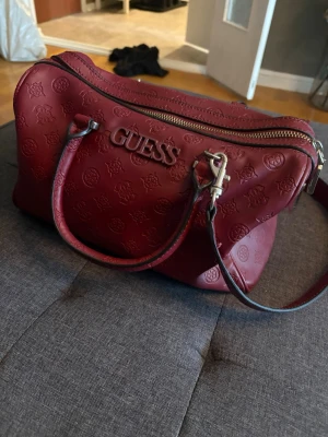 Vinröd handväska från Guess - Snygg vinröd handväska från Guess i präglat konstläder med logomönster över hela väskan. Väskan har både handtag och en justerbar axelrem samt silverfärgade detaljer och dragkedja. Perfekt storlek för det viktigaste och ger en trendig look.