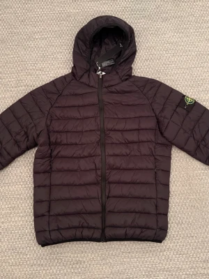 Svart pufferjacka från Stone Island - NY OANVÄND MED PRISTAGGEN KVAR! Snygg svart pufferjacka från Stone Island med huva och dragkedja framtill. Jackan har det klassiska Stone Island-märket på vänster ärm och quiltad design. Perfekt för kalla dagar och riktigt trendig stil. OBS! Det står att storleken är 3XL MEN den är som en XL eller som en stor LARGE skulle jag säga.