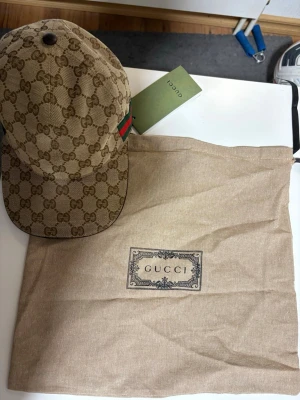 Beige keps från Gucci med GG-mönster - Säljer en beige keps från Gucci med det klassiska GG-monogrammönstret över hela kepsen. Kepsen har en grön och röd rand på sidan och justerbar rem bak. Materialet är canvas och insidan är fodrad i brunt tyg. Dustbag från Gucci medföljer. Storlek M
