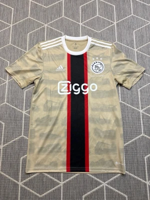 Ajax Adidas - Ajax fotbollströja i beige med svart och röd rand längs mitten, vita detaljer och Ziggo-logga på bröstet. Klassisk Ajax-logga med tre stjärnor och Adidas-märke. Tillverkad i lätt, ventilerande material och har korta ärmar samt vit krage.