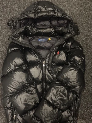 Svart dunjacka från Polo Ralph Lauren - (PRIS GÅR ATT DISKUTERAS) Snygg svart dunjacka från Polo Ralph Lauren med huva och dragkedja framtill. Jackan har en puffig design, två sidofickor med dragkedja och den klassiska röda loggan broderad på bröstet. Perfekt för kalla vinterdagar och riktigt varm. Jackan har ett litet hål på armen som jag har täckt över
