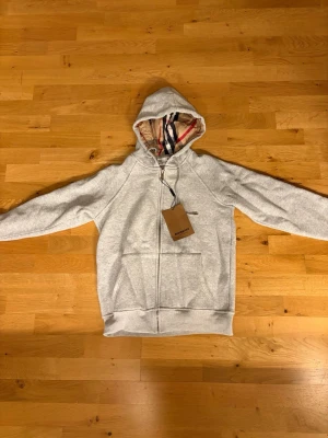 Grå zip hoodie från Burberry - Snygg grå zip hoodie från Burberry med klassiskt rutigt foder i huvan. Hoodien har dragkedja framtill, två stora fickor och justerbar dragsko i huvan. Tillverkad i mjukt material som känns skönt mot huden. Perfekt för en avslappnad och trendig look.