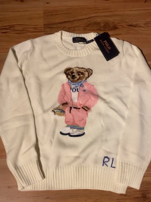 Vit stickad tröja Polo Ralph Lauren - Vit stickad tröja från Polo Ralph Lauren med klassisk björn broderad på framsidan i rosa outfit och blå scarf. Tröjan har rund hals, ribbade muddar och RL-broderi vid nederkanten. Perfekt för dig som gillar ikoniska detaljer och snygg design.