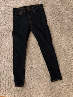 Mörkblå skinny jeans från Nudie - Säljer ett par mörkblå skinny jeans från Nudie Jeans med klassisk femficksmodell och kontrastsömmar. Jeansen har hög midja och är tillverkade i stretchig denim för en tight passform. Perfekta för dig som gillar en clean och stilren look.