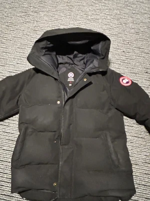 Svart dunjacka från Canada Goose macmillen - Snygg svart dunjacka från Canada Goose med huva och klassisk logotyp på ärmen. Jackan har dragkedja och tryckknappar framtill samt två stora fickor. Perfekt för kalla vinterdagar och riktigt varm tack vare dunfyllningen. Storleken är Xs men är dock stor i storleken så passar S också