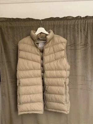 Beige dunväst från Ciszere - Stilren beige dunväst från Ciszere med hög krage och dragkedja framtill. Västen har två sidofickor med dragkedja och quiltad design. Insidan har en rolig tvättinstruktionslapp med tecknade figurer. Perfekt för lager-på-lager och enkel att matcha.