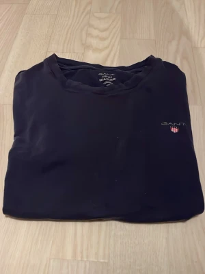 Marinblå t-shirt från GANT med logga - En svart t-shirt från GANT med liten broderad logga och sköld på bröstet. Klassisk rund halsringning och enkel design som passar till det mesta. Tillverkad i mjuk bomull för en skön känsla.