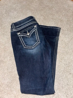 Mörkblå miss me bootcut jeans - Miss me mörkblå bootcut jeans i storlek 31 med vita kontrastsömmar och detaljerade bakfickor med en dekorativ knapp. Bra skick förutom skav vid slutet av benen då de har släpat lite i marken annars inget man tänker på! Säljer då de är för stora för mig❣️Köpta för 2200 säljer för 1000🥰