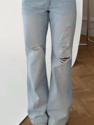 Slutsålda bootcut jeans - Säljer ett par ljusblåa bootcut jeans med flera slitningar och hål på benen. Jeansen har en avslappnad passform och är tillverkade i denim. Perfekta för en trendig och avslappnad stil.