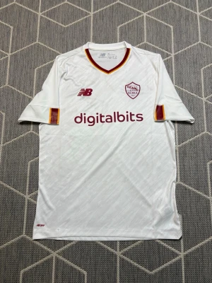 AS Roma New Balance - Snygg vit AS Roma matchtröja från New Balance med klubbmärke och sponsortryck i vinrött. Tröjan har korta ärmar, V-ringning med gul och vinröd kant samt ventilerande material perfekt för fotbollsträning.