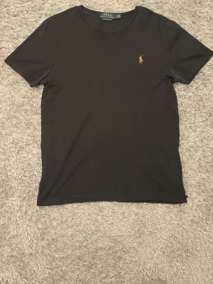 Svart t-shirt från Polo Ralph Lauren - Klassisk svart t-shirt från Polo Ralph Lauren med gul broderad logga på bröstet. T-shirten har rund halsringning och korta ärmar. Tillverkad i mjuk bomull för en skön känsla och enkel stil.