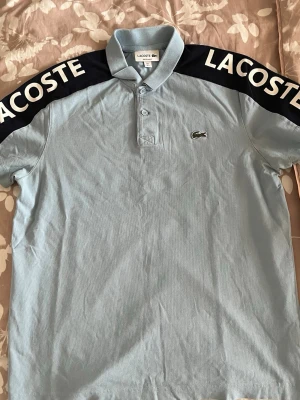 Ljusblå Lacoste t-shirt med logga - Snygg ljusblå t-shirt från Lacoste med klassisk krage och knappar framtill. Svarta partier över axlarna med stor vit Lacoste-text. Ikoniska krokodilloggan på bröstet. Tillverkad i mjuk bomull, perfekt för en avslappnad och sportig stil.
