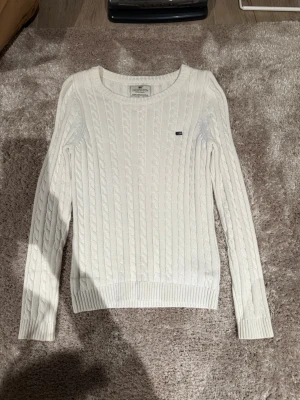 Vit kabelstickad tröja Lexington - Kabelstickad vit tröja från Lexington med rund halsringning och diskret logga på bröstet. Tröjan har långa ärmar och ribbade muddar vid ärmslut och nederkant. Perfekt för en klassisk och stilren look.