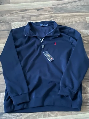 Mörkblå halvzip från Polo Ralph Lauren - Snygg mörkblå tröja med halv dragkedja från Polo Ralph Lauren. Klassisk design med röd broderad logga på bröstet och ribbade muddar. Perfekt för en stilren och avslappnad look.