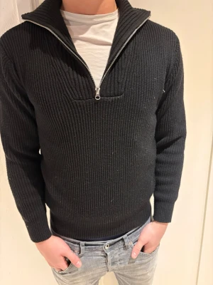 Svart stickad tröja med halv dragkedja - Säljer en svart ribbstickad tröja med halv dragkedja och hög krage. Tröjan har långa ärmar och en klassisk, stilren look som passar till det mesta. Perfekt för lager-på-lager och enkel att matcha med jeans eller chinos.