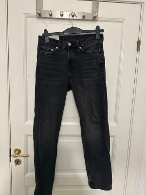 Svarta raka jeans från H&M - Säljer ett par svarta jeans med rak passform och klassisk femficksdesign från H&M. Jeansen har knappgylf och silverfärgade nitar. Materialet är denim i bomull och färgen har en lätt tvättad look för en cool vibe. Storlek 29/32