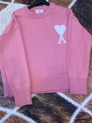 Rosa stickad tröja från Ami Paris - Säljer en rosa stickad tröja från Ami Paris med ett vitt hjärta och bokstaven 'A' på bröstet. Tröjan har rund halsringning, långa ärmar och breda ribbade muddar vid ärmslut och nederkant. Perfekt för dig som gillar trendiga och bekväma plagg.