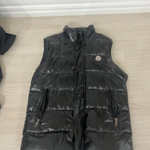 Svart dunväst från Moncler - Snygg svart dunväst från Moncler med glansig finish och hög krage. Västen har två dragkedjeförsedda fickor framtill och Moncler-logga på bröstet. Perfekt för lager på lager och ger en cool streetwear-look.
