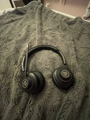 Svarta over-ear hörlurar JLab - Snygga svarta over-ear hörlurar från JLab med rundad design och vadderad bygel. Hörlurarna har en utfällbar mikrofon och mjuka öronkuddar i konstläder för extra komfort. Perfekta för gaming, musik eller videosamtal.