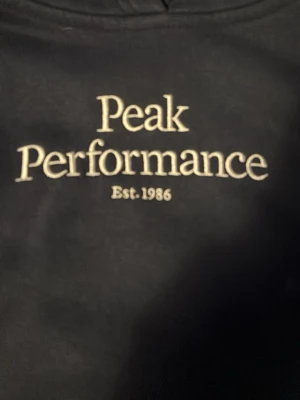 Svart hoodie Peak Performance - Säljer en svart hoodie från Peak Performance med vit broderad logga och texten 'Est. 1986' på bröstet. Klassisk modell med huva och mjukt material, perfekt för en avslappnad stil. Passar dig som gillar stilrena och sportiga plagg.