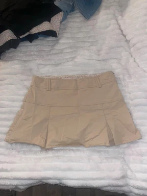 Beige kortkjol från att. - Snygg beige kortkjol från att. med veckade detaljer och bälteshällor i midjan. Kjolen har ett blommigt foder på insidan som ger en söt touch. Perfekt för dig som gillar stilrena och feminina plagg.