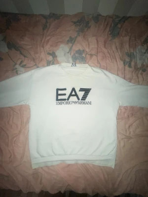 Vit EA7 (Emporio Armani) M/S - Säljer en stilren vit sweatshirt från Emporio Armani EA7 med stor svart logga på bröstet. Tröjan har rund halsringning, långa ärmar och ribbade muddar vid ärmslut och nederkant. Perfekt för en clean och trendig look.