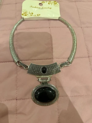 Silvrigt halsband med svart sten - Unikt halsband i silvrig metall med en stor oval svart sten som hänge och detaljerad dekor ovanför. Halsbandet har en kraftig, böjd kedja och antika mönster på metallen. Perfekt för dig som gillar statement-smycken och vill sticka ut.