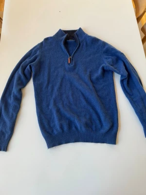 Blå halvzip tröja från James Pringle - Mysig blå stickad tröja från James Pringle med halvzip och ribbade muddar. Tröjan har hög krage och är tillverkad i mjukt material, perfekt för lager på lager. Skriv ifall du har några frågor!