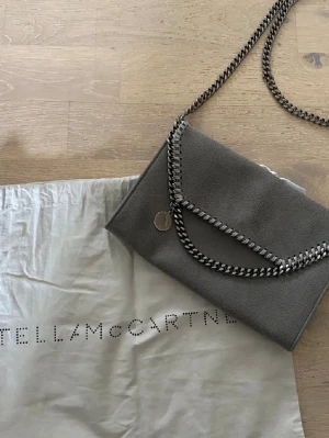 Grå kuvertväska Stella McCartney - Snygg grå kuvertväska från Stella McCartney med silverfärgad kedja och dekorativa kedjedetaljer längs kanten. Har bara använt två gånger och är i superfint skick!! Köpt i butik för ett tag sedan. Har ej kvar kvitto men har dustbag