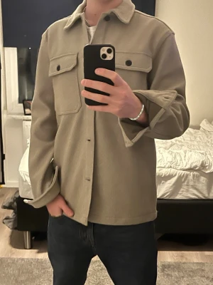 Beige overshirt med fickor - Stilren beige overshirt med klassisk krage och två stora bröstfickor med lock. Overshirten har raka ärmar och knäppning framtill. Perfekt att bära öppen eller stängd för en avslappnad look. Materialet ser ut att vara mjukt och lite tjockare, troligen ullblandning.
