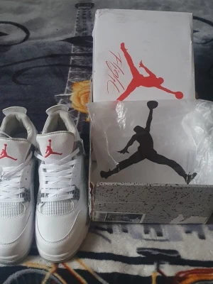Air Jordan 4 Retro White/Red - Snygga Air Jordan 4 Retro sneakers i vitt läder med röda detaljer och klassisk Jumpman-logga på plösen. Skorna har meshpaneler, gråa accenter och en robust sula. Perfekta för dig som gillar streetwear och vill sticka ut med en ikonisk modell.