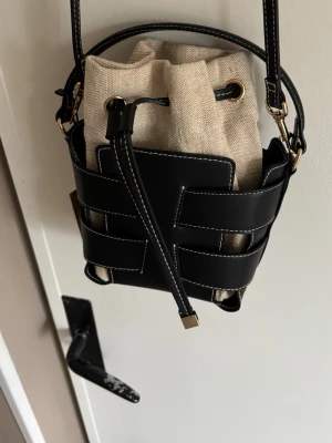 Bucket bag  - Säljer en trendig svart bucket bag med vita sömmar och detaljer i guld. Väskan har en beige canvasinsats med dragsko och justerbar axelrem. Snygg kombination av läderimitation och tyg som ger en modern look.