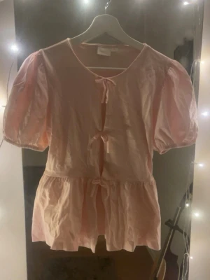 Ljusrosa blus med puffärm och rosetter - Aldrig använd! Supersöt ljusrosa blus med puffiga korta ärmar och tre rosetter framtill. Blusen har en luftig passform och volang nedtill,  Tillverkad i mjukt bomullstyg och passar perfekt till både jeans och kjol.