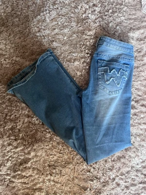Blå jeans från Wrangler med broderi - Säljer ett par klassiska blå jeans från Gina med ljus tvätt och snygga broderade detaljer i form av ett W på bakfickorna. Jeansen är bootcut  och är tillverkade i slitstark denim. Perfekta för en avslappnad och trendig look. Säljer då de är för stora så kan tänka mig att byta mot ett par lika dana i S🤍