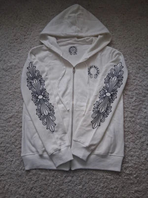 Vit hoodie med svart tryck - Säljer en vit hoodie från Chrome Hearts med dragkedja och stora svarta grafiska tryck längs båda ärmarna samt på bröstet och ryggen. Hoodien har huva och ribbade muddar. Materialet är mjukt och skönt bomullstyg.