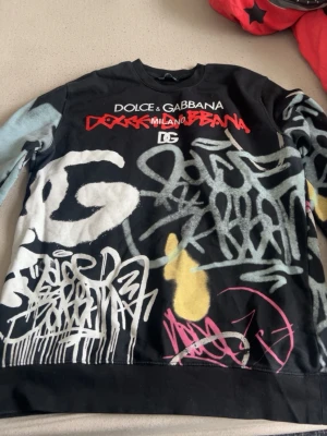 Dolce & Gabbana graffiti hoodie - Säljer en svart hoodie från Dolce & Gabbana med streetinspirerad graffiti-design i vitt, grått, rött, gult och rosa. Tröjan har tryckta loggor och text på bröstet samt färgstarka detaljer över hela plagget. Perfekt för dig som gillar unik och edgy stil.